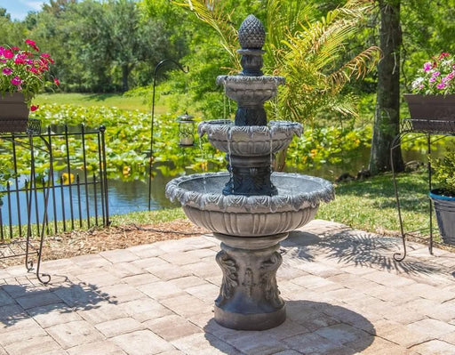 Stone fountain in a garden setting with flowers and a pond in the background