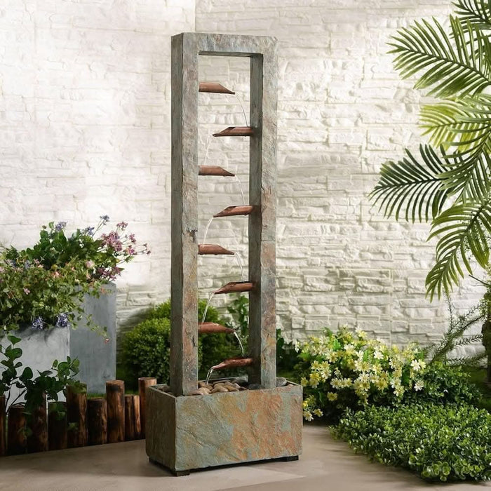Slate water fountain with copper spouts in a stone base, surrounded by garden plants.