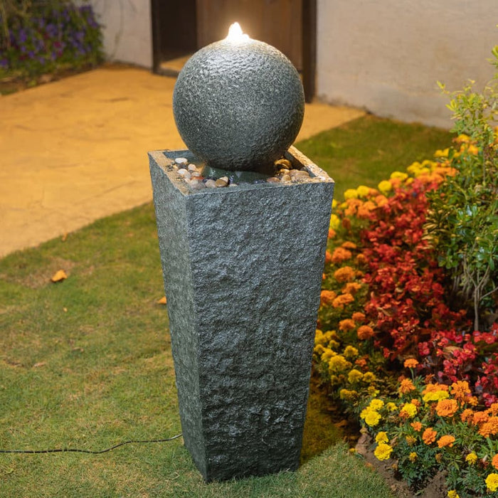 A tower & ball water feature near a flower bed.