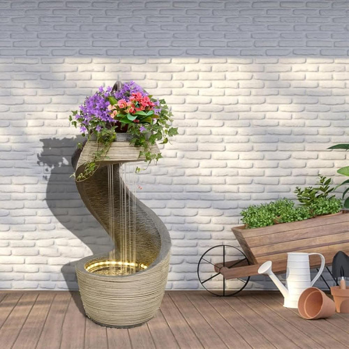 Modern spiral water fountain with LED lighting and a vibrant flower arrangement on top, set against a white brick wall with garden tools nearby.