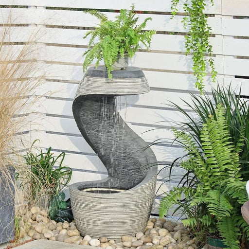 Curved outdoor water fountain with a ribbed stone finish and built-in planter holding a fern, set among garden pebbles and greenery.