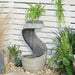 Curved outdoor water fountain with a ribbed stone finish and built-in planter holding a fern, set among garden pebbles and greenery.