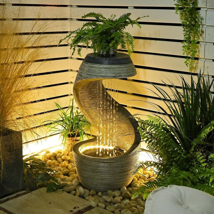 Curved outdoor water fountain with warm LED lighting and a fern planter on top, set among garden pebbles and greenery in a softly lit corner.