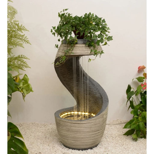 Indoor water fountain with a curved spiral design, warm LED lighting, and a cascading ivy plant on top, placed on a light pebble surface.