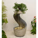 Indoor water fountain with a curved spiral design, warm LED lighting, and a cascading ivy plant on top, placed on a light pebble surface.