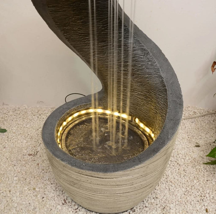 Close-up of a modern spiral water fountain with cascading streams and soft LED lighting around the basin, set on a white pebble base.