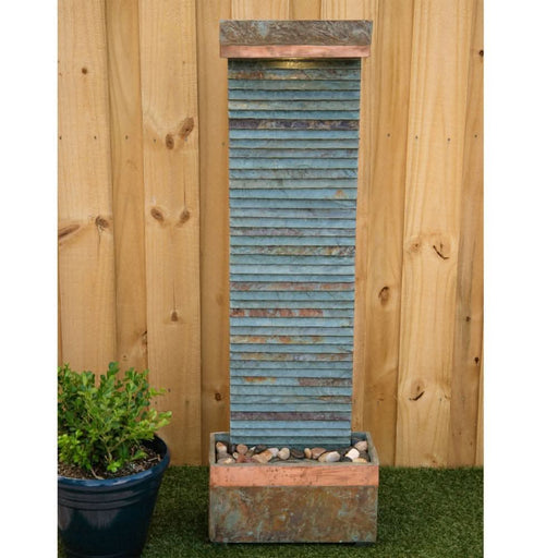 Tall slate water feature with horizontal ridges and copper trim, placed in a garden setting with pebbles at the base.