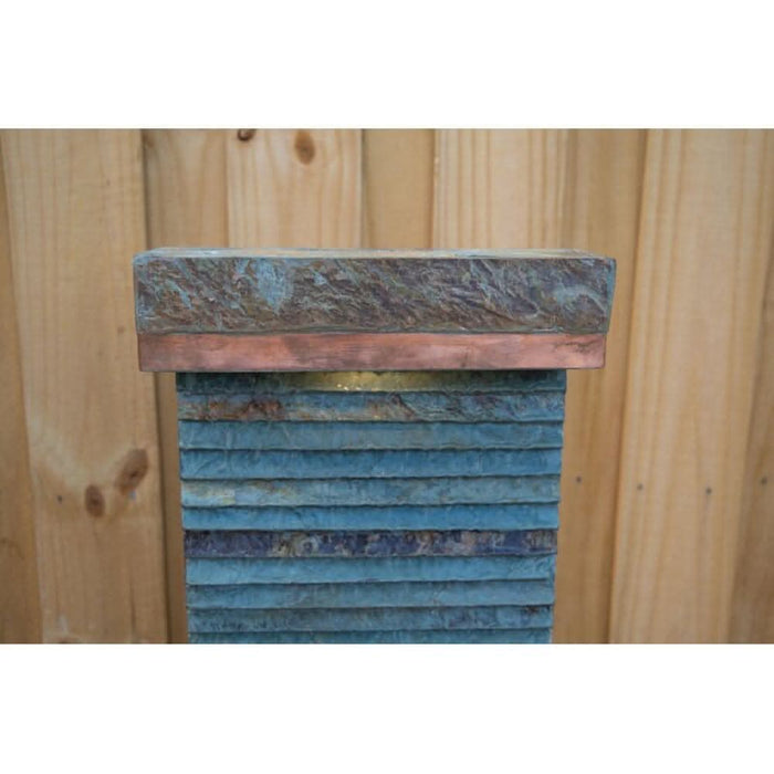 Top section of a slate water feature with copper edging and layered stone texture, set against a timber fence.