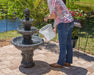 Filling three-tier outdoor water fountain with a bucket on a sunny patio beside a garden pond.