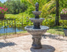 Stone fountain in a garden setting with flowers and a pond in the background