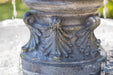 Close-up of ornate acanthus leaf detailing on the pedestal base of a weathered stone water fountain.