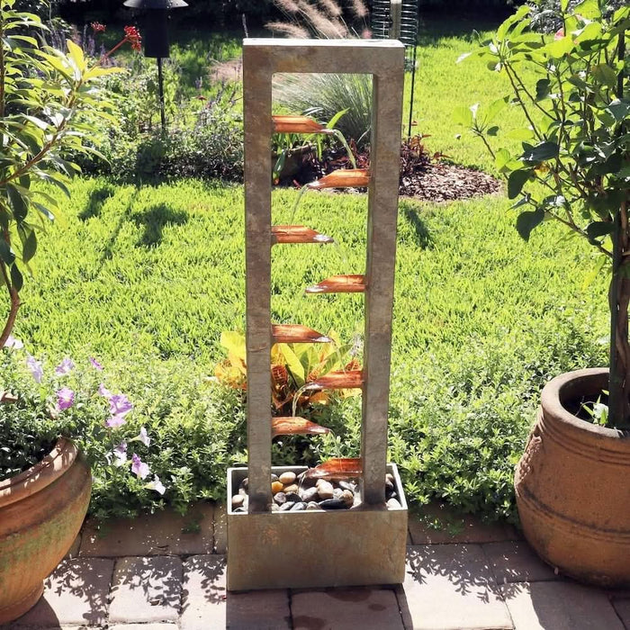 Outdoor slate water fountain with copper spouts cascading into a stone-filled base, set in a garden.